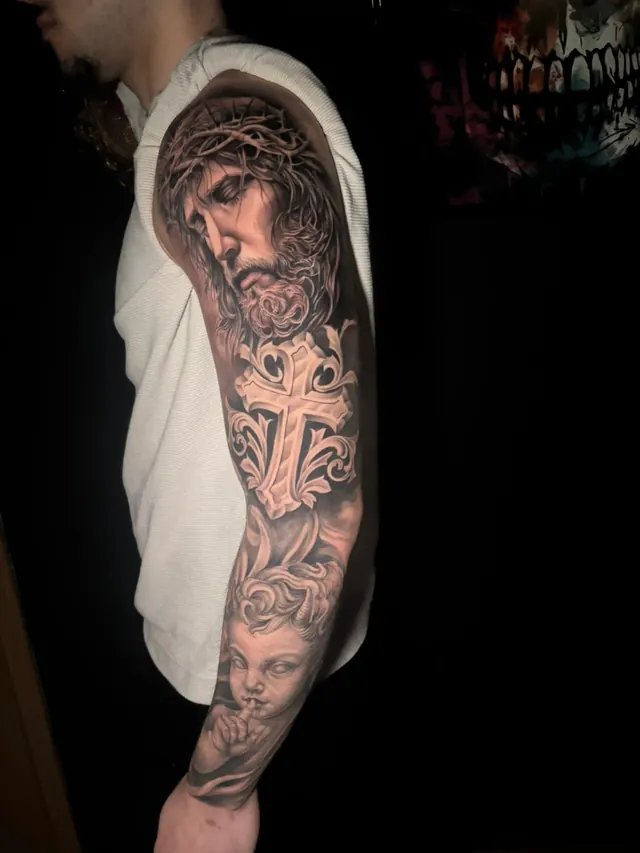 Black and grey religious realism sleeve tattoo with Christ, an ornate cross and a cherub