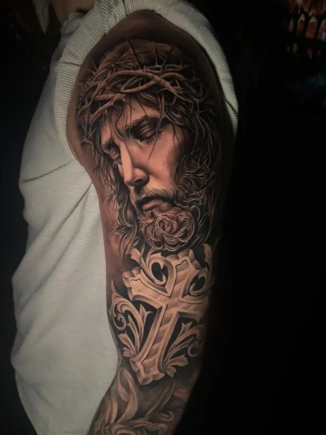 Religious realism upper-arm tattoo of Christ with a crown of thorns above an ornate cross